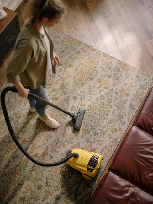 A woman using a yellow vacuum cleaner with a black hose to deep clean a patterned cream and orange rug in a living room. The rug covers a wooden floor, and a red leather sofa is partially visible to the right. The room is well-lit, highlighting the surface cleanliness achieved through professional domestic cleaning services by Carpet Cleaning Edgware. The woman is wearing a beige jacket, jeans, and white sneakers, focusing on the task of surface cleaning to maintain hygiene and freshness in the residential space.