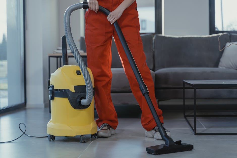 A woman with curly hair, wearing a beige jacket and blue jeans, is engaged in surface cleaning with a vacuum cleaner powered by a yellow canister, cleaning a patterned area rug in a living room. The r