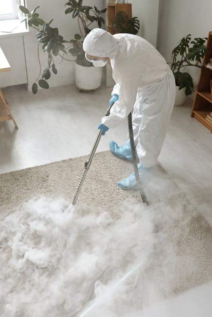Close-up image of a person vacuuming a patterned area rug with a black upright vacuum cleaner, which is positioned on the carpet and operated by someone wearing white sneakers and dark pants. The room