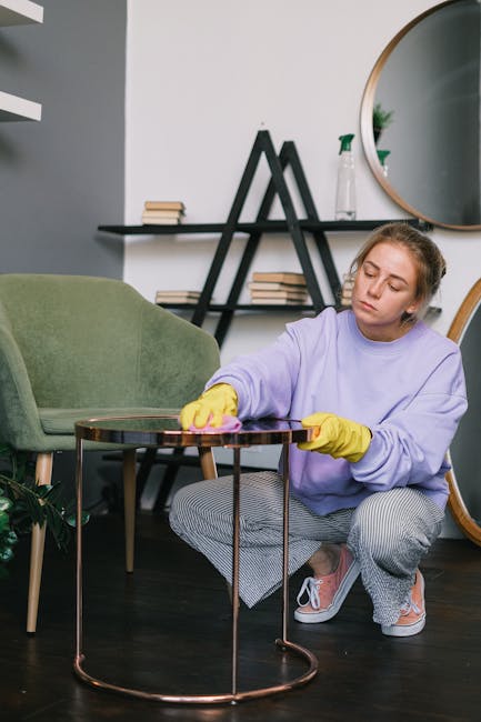 A close-up of a person's hand holding a handheld upholstery cleaning tool connected to a vacuum or extraction device, actively cleaning a beige fabric upholstered chair or sofa. The fabric on the seat