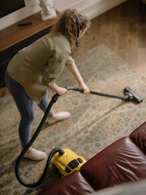 A woman with curly hair and casual clothing is engaged in surface cleaning using a yellow vacuum cleaner with a long hose attachment. She is standing on a patterned area rug in a well-lit, cozy living