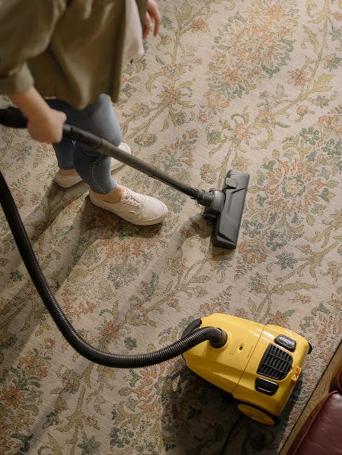 A woman with curly hair and casual clothing is engaged in surface cleaning using a yellow vacuum cleaner with a long hose attachment. She is standing on a patterned area rug in a well-lit, cozy living